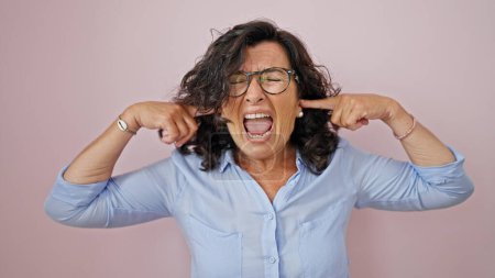 Téléchargez les photos : Moyen Age femme hispanique criant bruyamment couvrant les oreilles avec les doigts sur fond rose isolé - en image libre de droit