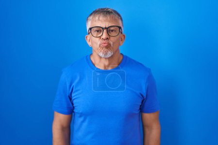 Foto de Hombre hispano con el pelo gris de pie sobre fondo azul mirando a la cámara soplando un beso en el aire siendo encantador y sexy. expresión de amor. - Imagen libre de derechos