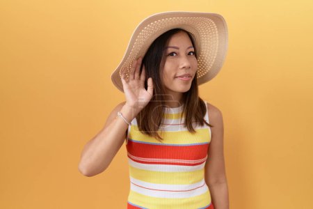Photo for Middle age chinese woman wearing summer hat over yellow background smiling with hand over ear listening an hearing to rumor or gossip. deafness concept. - Royalty Free Image