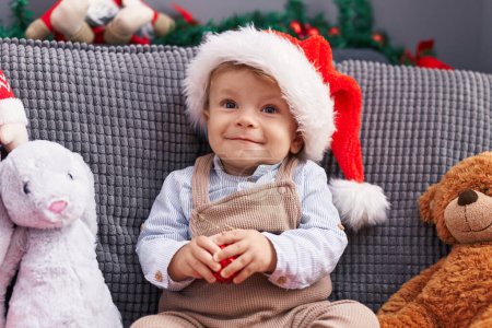 Photo for Adorable blond toddler sitting on sofa wearing christmas hat at home - Royalty Free Image
