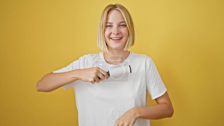 Téléchargez les photos : Jeune femme blonde utilisant un rouleau de bâton pour nettoyer les poils sur fond jaune isolé - en image libre de droit