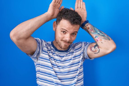 Photo for Young hispanic man standing over blue background doing bunny ears gesture with hands palms looking cynical and skeptical. easter rabbit concept. - Royalty Free Image