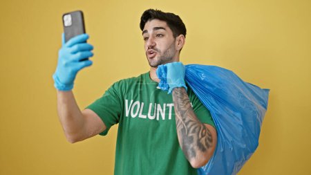 Foto de Joven hispano voluntario sosteniendo bolsa de basura teniendo videollamada sobre aislado fondo amarillo - Imagen libre de derechos
