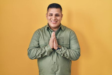 Téléchargez les photos : Jeune homme hispanique debout sur fond jaune priant avec les mains ensemble demandant pardon souriant confiant. - en image libre de droit