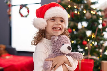 Foto de Adorable chica rubia abrazando oso de peluche de pie junto al árbol de Navidad en casa - Imagen libre de derechos