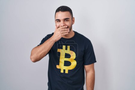 Foto de Joven hombre hispano vistiendo camiseta bitcoin riendo y riéndose avergonzado cubriendo la boca con las manos, chismes y concepto de escándalo - Imagen libre de derechos