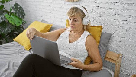 Photo for Relaxed middle age blonde woman enjoying morning tunes, beautifully sitting in her bedroom, laptop on her lap, headphones on, her serious face glowing in the soft indoor light. - Royalty Free Image