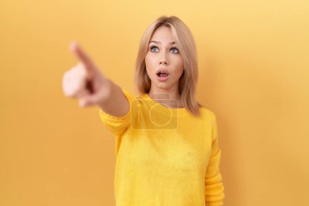 Photo for Young caucasian woman wearing yellow sweater pointing with finger surprised ahead, open mouth amazed expression, something on the front - Royalty Free Image