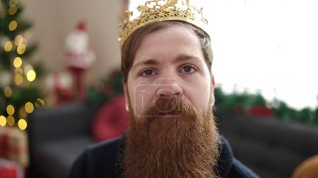 Photo for Young redhead man wearing king crown sitting on sofa by christmas tree at home - Royalty Free Image