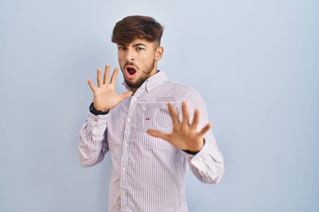 Photo for Arab man with beard standing over blue background afraid and terrified with fear expression stop gesture with hands, shouting in shock. panic concept. - Royalty Free Image
