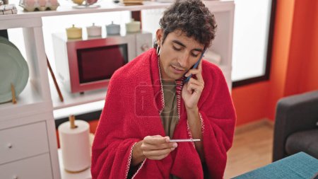 Photo for Young hispanic man talking on smartphone looking thermometer at dinning room - Royalty Free Image