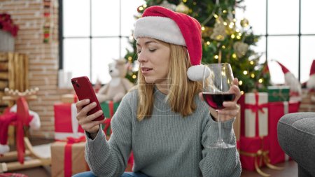 Foto de Mujer rubia joven usando smartphone bebiendo vino celebrando la Navidad en casa - Imagen libre de derechos