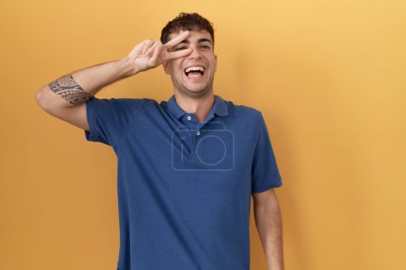 Photo for Young hispanic man standing over yellow background doing peace symbol with fingers over face, smiling cheerful showing victory - Royalty Free Image
