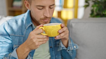 Photo for Young caucasian man smelling cup of coffee sitting on sofa at home - Royalty Free Image
