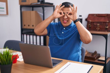 Foto de Un joven hispano trabajando en la oficina con un portátil haciendo un buen gesto como binoculares sacando la lengua, ojos mirando a través de los dedos. expresión loca. - Imagen libre de derechos