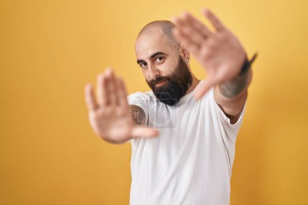 Foto de Joven hombre hispano con barba y tatuajes de pie sobre fondo amarillo haciendo marco usando las manos palmas y los dedos, perspectiva de la cámara - Imagen libre de derechos