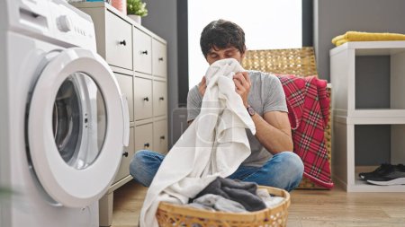 Photo for Young hispanic man washing clothes smelling clean towel at laundry room - Royalty Free Image