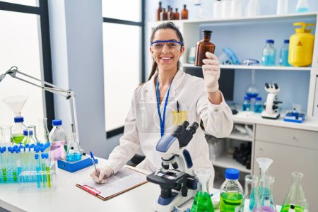 Photo for Young beautiful hispanic woman scientist writing report holding bottle at laboratory - Royalty Free Image