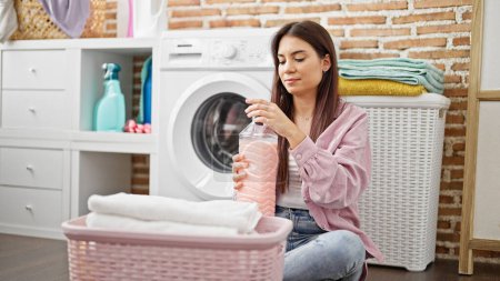 Foto de Joven hermosa mujer hispana lavando ropa sosteniendo botella de detergente en la sala de lavandería - Imagen libre de derechos
