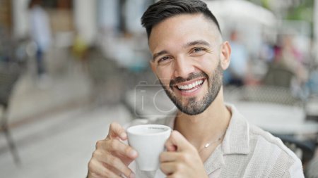 Téléchargez les photos : Jeune homme hispanique souriant boire du café en toute confiance à la terrasse du café - en image libre de droit