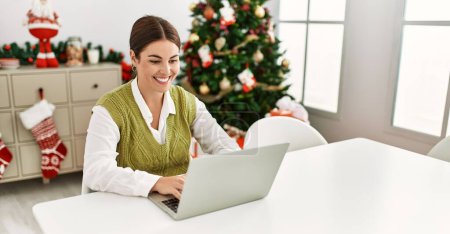 Photo for Young beautiful hispanic woman using laptop sitting by christmas tree at home - Royalty Free Image