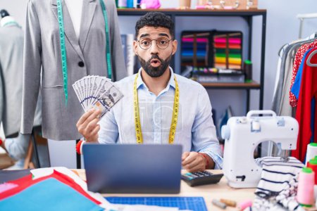 Photo for Hispanic man with beard dressmaker designer holding dollars scared and amazed with open mouth for surprise, disbelief face - Royalty Free Image