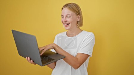 Téléchargez les photos : Jeune femme blonde utilisant un ordinateur portable souriant sur fond jaune isolé - en image libre de droit