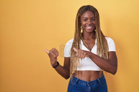 Foto de Mujer afroamericana con el pelo trenzado de pie sobre fondo amarillo apuntando a la espalda con la mano y los pulgares hacia arriba, sonriendo confiado - Imagen libre de derechos