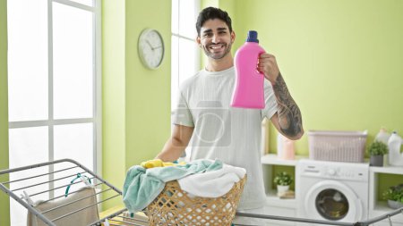 Foto de Joven hispano sosteniendo botella de detergente junto al tendedero en la lavandería - Imagen libre de derechos
