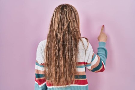 Photo for Young blonde woman standing over pink background posing backwards pointing ahead with finger hand - Royalty Free Image