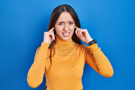 Photo for Young brunette woman standing over blue background covering ears with fingers with annoyed expression for the noise of loud music. deaf concept. - Royalty Free Image