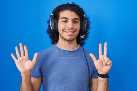 Téléchargez les photos : Homme hispanique aux cheveux bouclés écoutant de la musique à l'aide d'écouteurs montrant et pointant vers le haut avec les doigts numéro huit tout en souriant confiant et heureux. - en image libre de droit