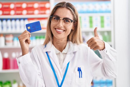 Téléchargez les photos : Jeune femme blonde travaillant à la pharmacie pharmacie tenant la carte de crédit souriant heureux et positif, pouce levé faire excellent et signe d'approbation - en image libre de droit