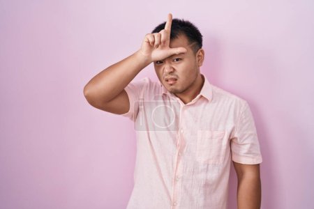 Photo for Chinese young man standing over pink background making fun of people with fingers on forehead doing loser gesture mocking and insulting. - Royalty Free Image
