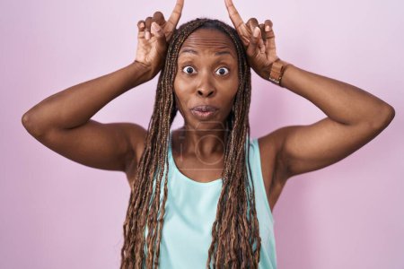 Téléchargez les photos : Afro-américaine femme debout sur fond rose faire geste drôle avec doigt sur la tête comme cornes de taureau - en image libre de droit