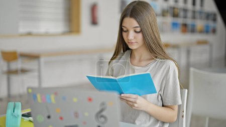 Téléchargez les photos : Jeune belle fille étudiant livre de lecture en utilisant un ordinateur portable à la bibliothèque - en image libre de droit