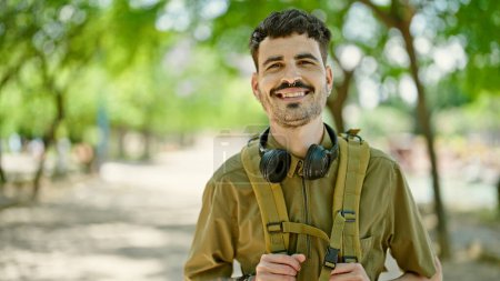Téléchargez les photos : Jeune homme hispanique touriste portant sac à dos et écouteurs souriant au parc - en image libre de droit