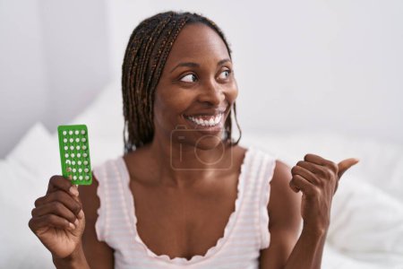 Photo for African american woman holding birth control pills pointing thumb up to the side smiling happy with open mouth - Royalty Free Image