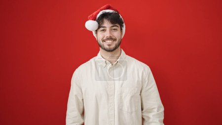 Téléchargez les photos : Jeune homme hispanique souriant confiant portant un chapeau de Noël sur fond rouge isolé - en image libre de droit