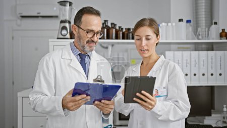 Photo for Two scientists engrossed in reading an intriguing document on clipboard at the lab, speaking about their startling discovery, while working on a touchpad together. - Royalty Free Image