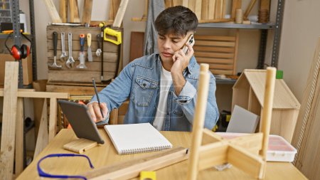 Foto de Experiencia de carpintero hispano joven multitarea, hablando en un teléfono inteligente mientras se utiliza un touchpad en un taller de carpintería ocupado - Imagen libre de derechos