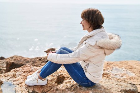 Foto de Mujer de mediana edad sonriendo confiado sentado en la roca a la orilla del mar - Imagen libre de derechos