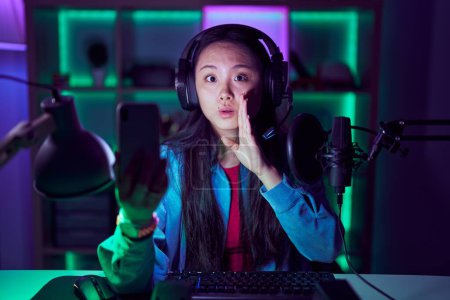 Téléchargez les photos : Jeune femme asiatique jouer à des jeux vidéo avec smartphone main sur la bouche racontant rumeur secrète, chuchotant conversation malicieuse - en image libre de droit