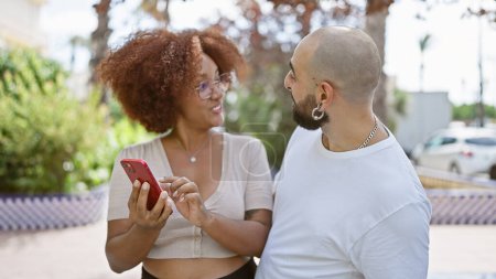 Photo for Confident, happy interracial couple smiling together, standing in sunlit park with their smartphone, effortlessly sharing love and laughter outdoors - Royalty Free Image