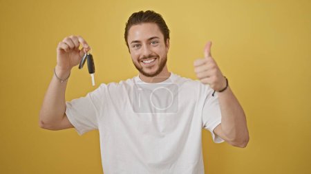 Foto de Joven hispano sosteniendo la llave de un auto nuevo haciendo un gesto con el pulgar hacia arriba sobre un fondo amarillo aislado - Imagen libre de derechos