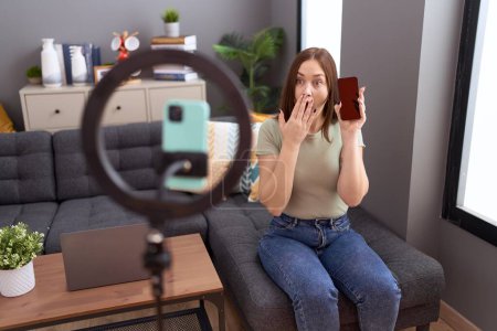 Photo for Beautiful brunette woman recording vlog tutorial with smartphone at home covering mouth with hand, shocked and afraid for mistake. surprised expression - Royalty Free Image