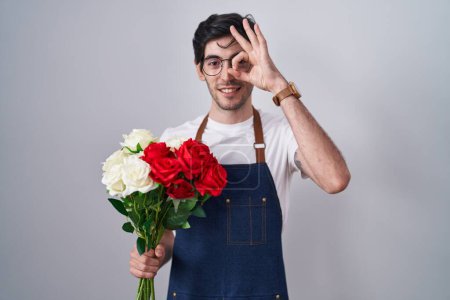 Téléchargez les photos : Jeune homme hispanique tenant un bouquet de roses blanches et rouges faisant un geste correct avec la main souriante, les yeux regardant à travers les doigts avec un visage heureux. - en image libre de droit
