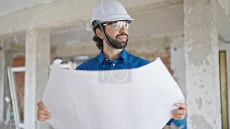 Photo for Young hispanic man architect holding blueprints looking around at construction site - Royalty Free Image