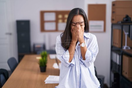 Junge hispanische Frau im Büro reibt sich die Augen vor Müdigkeit und Kopfschmerzen, schläfriger und müder Miene. Sehschwäche 