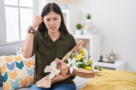 Photo for Chinese young woman holding bouquet of white flowers angry and mad raising fist frustrated and furious while shouting with anger. rage and aggressive concept. - Royalty Free Image